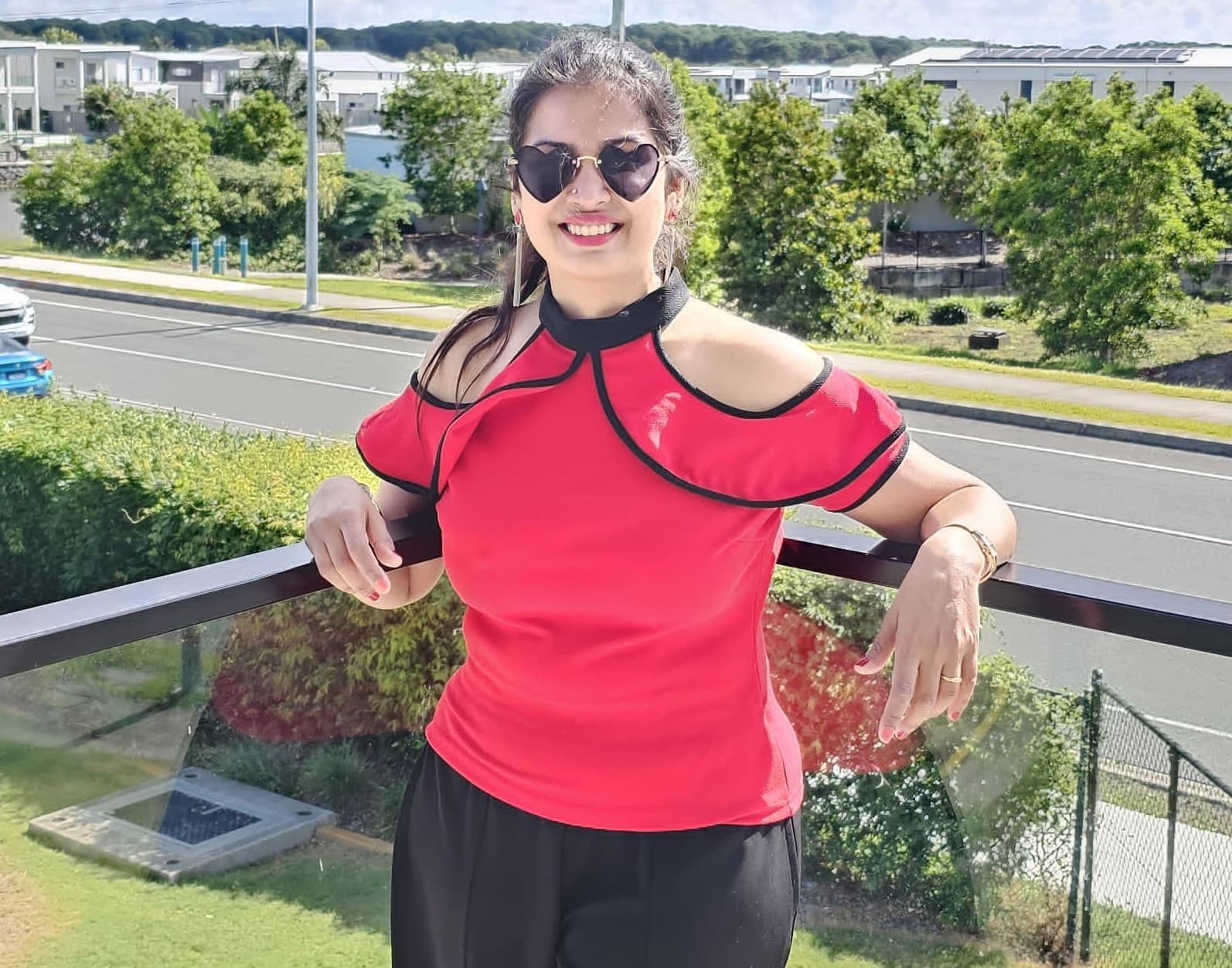 Smiling woman wearing a red cold-shoulder top and heart-shaped sunglasses on an outdoor balcony.